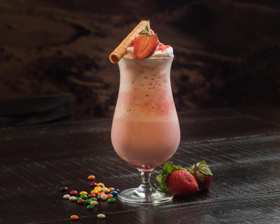 Hyatt Regency Dubai Creek Heights Hotel Exterior photo The photo shows a tall, elegant glass filled with a creamy, pink beverage that looks like a strawberry smoothie or milkshake. The drink is topped with whipped cream, a slice of strawberry, and a decorative wafer stick. Next to the glass, there are fr