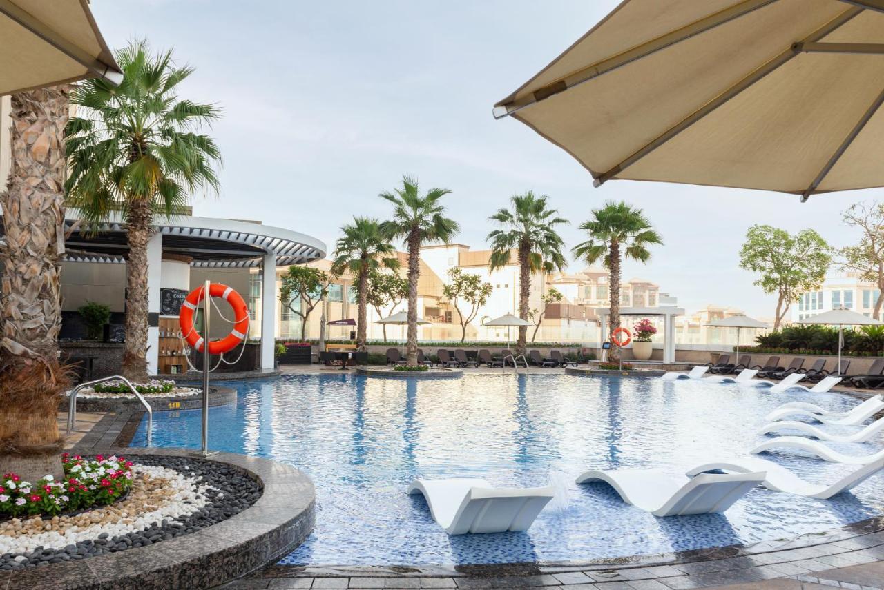 Hyatt Regency Dubai Creek Heights Hotel Exterior photo The photo showcases a serene swimming pool area surrounded by palm trees. The pool features a smooth, blue surface and has several lounge chairs partially submerged in the water. A large umbrella provides shade for the lounging area. In the backgroun