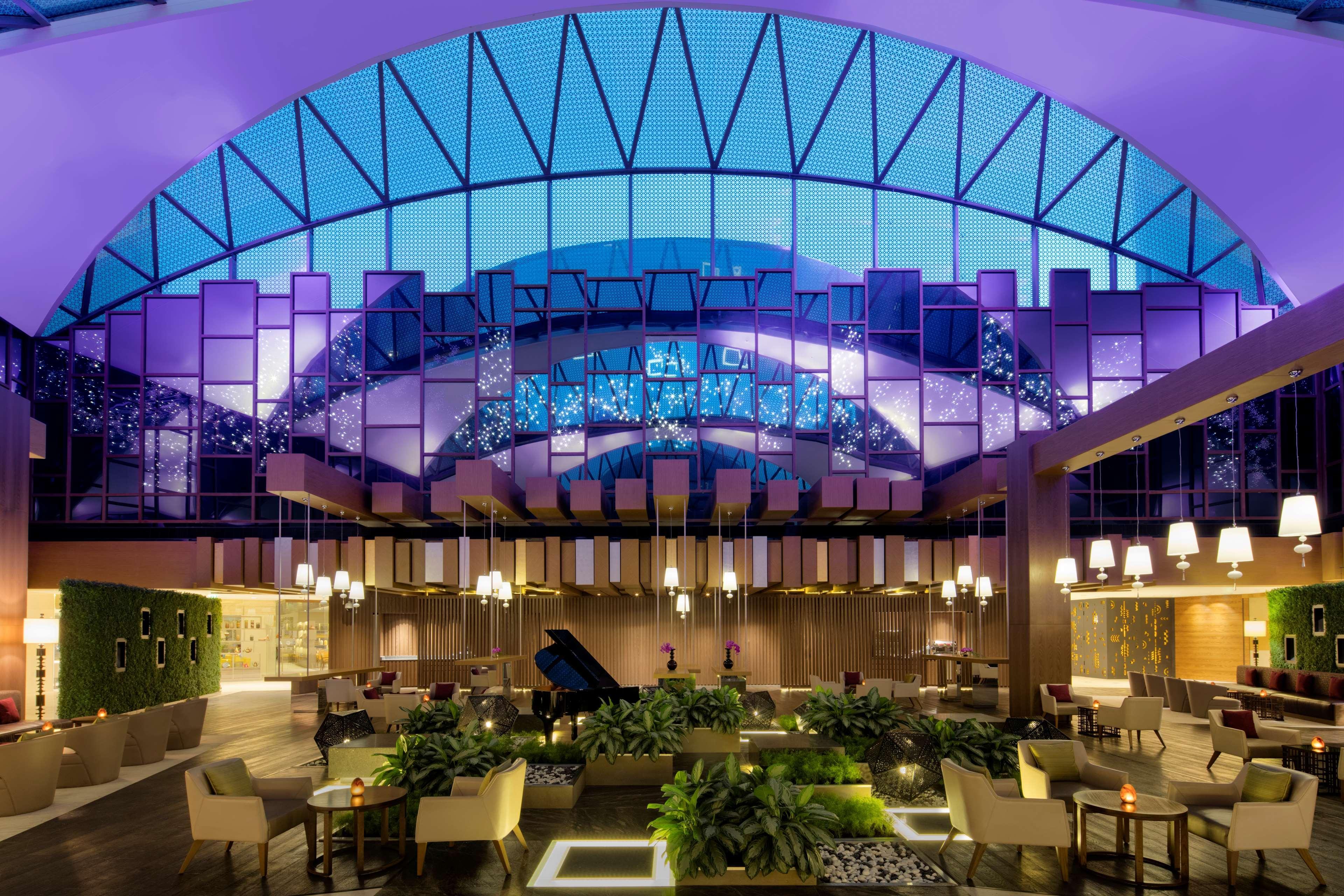 Hyatt Regency Dubai Creek Heights Hotel Exterior photo The photo showcases a modern, stylish interior of a large atrium or lounge area. The space features a high ceiling with a striking geometric design and is illuminated by vibrant purple and blue lighting, creating a dramatic atmosphere. There are nume