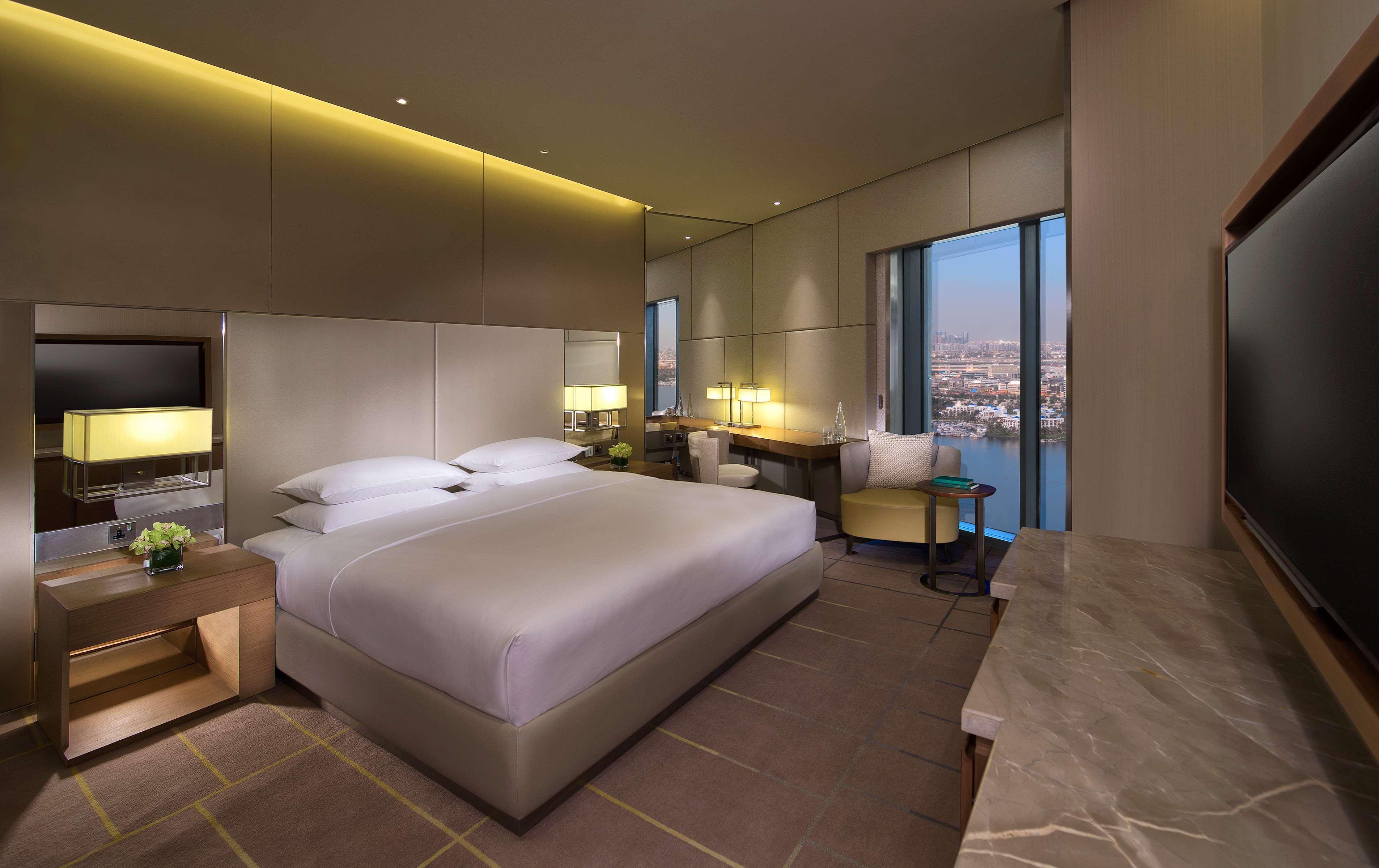 Hyatt Regency Dubai Creek Heights Hotel Exterior photo The image shows a modern hotel room. It features a large bed with white linens positioned against a wall. On either side of the bed, there are stylish lamps and small tables. A flat-screen television is mounted on the wall opposite the bed. The room 