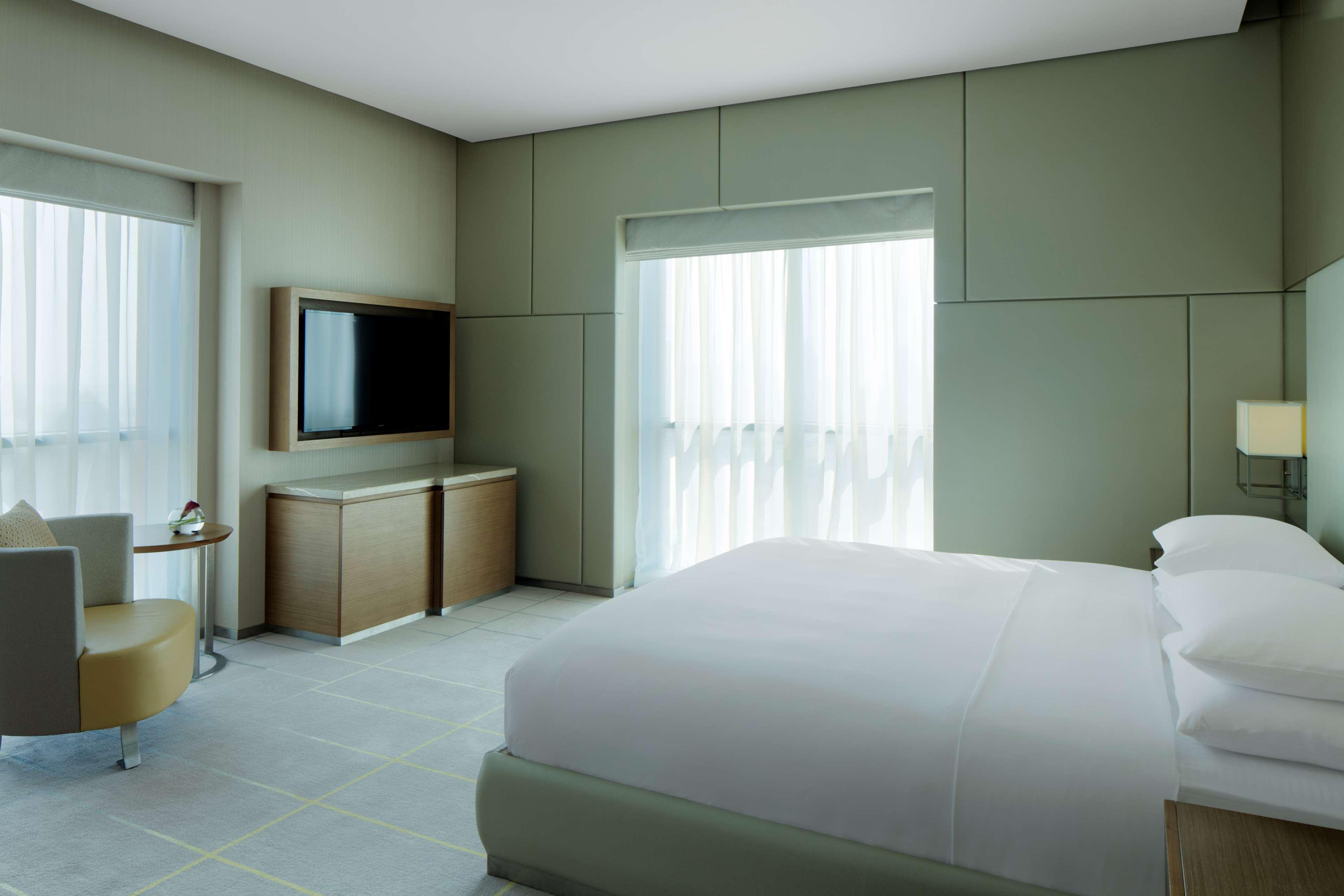 Hyatt Regency Dubai Creek Heights Hotel Exterior photo The photo shows a modern hotel room. It features a neatly made bed with white linens, a minimalist style. There is a medium-sized television mounted on the wall opposite the bed. A set of light-colored curtains allows natural light to enter through a